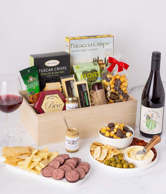 Gourmet gift box featuring a variety of snacks including focaccia crisps, Tuscan crisps, brie cheese, almonds, olives, mustard, popcorn, and a bottle of Hill &amp; Harbor Pinot Noir, with a glass of red wine and an assortment of crackers, sausage, and cheese 