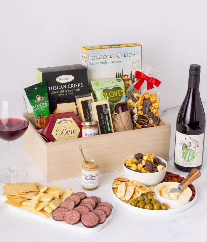 Gourmet gift box featuring a variety of snacks including focaccia crisps, Tuscan crisps, brie cheese, almonds, olives, mustard, popcorn, and a bottle of Hill & Harbor Pinot Noir, with a glass of red wine and an assortment of crackers, sausage, and cheese 