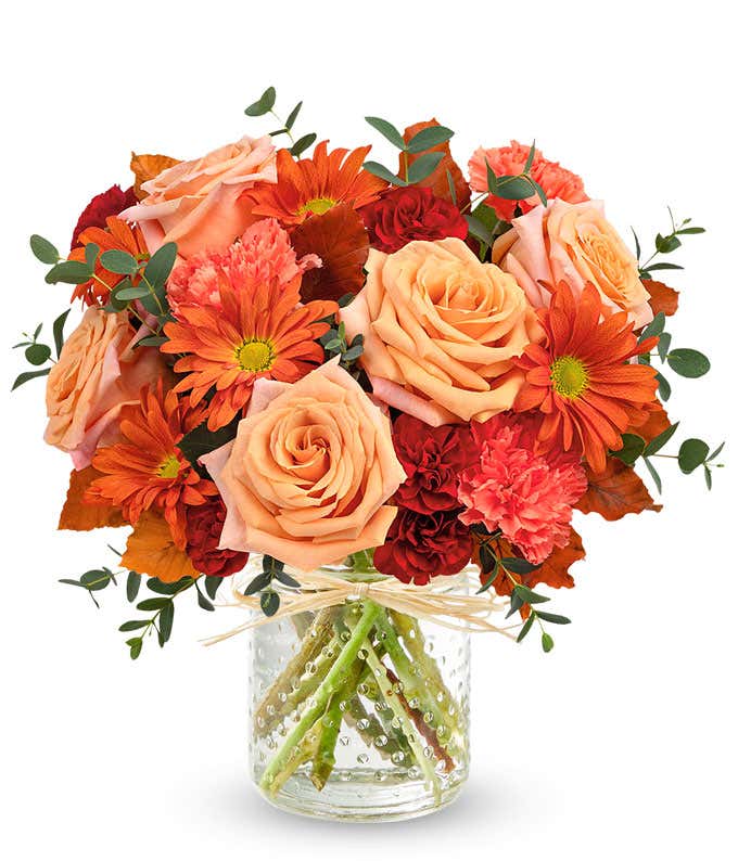 Light Orange Roses, Orange Carnations, Maroon mini carnations, bronze daisy spray chrysanthemums, brown copper beech, and parvifolia eucalyptus in a copper cylinder vase with patina against a white background